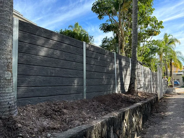 Dark timber-look sleeper retaining wall beside a driveway and palm trees